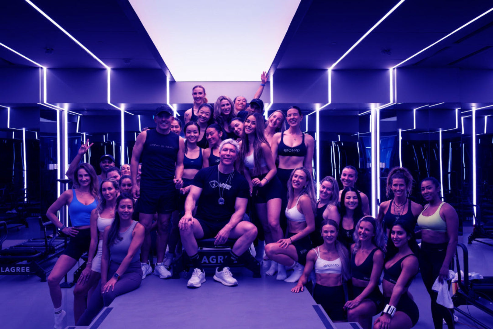 Group of people posing together in a Lagree fitness class with mirrors, fitness equipment, and Sebastian Lagree